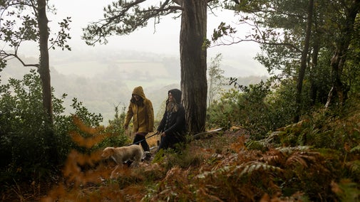 Two people walking a dog in the rain. They are walking along a wooded path with a misty view behind them